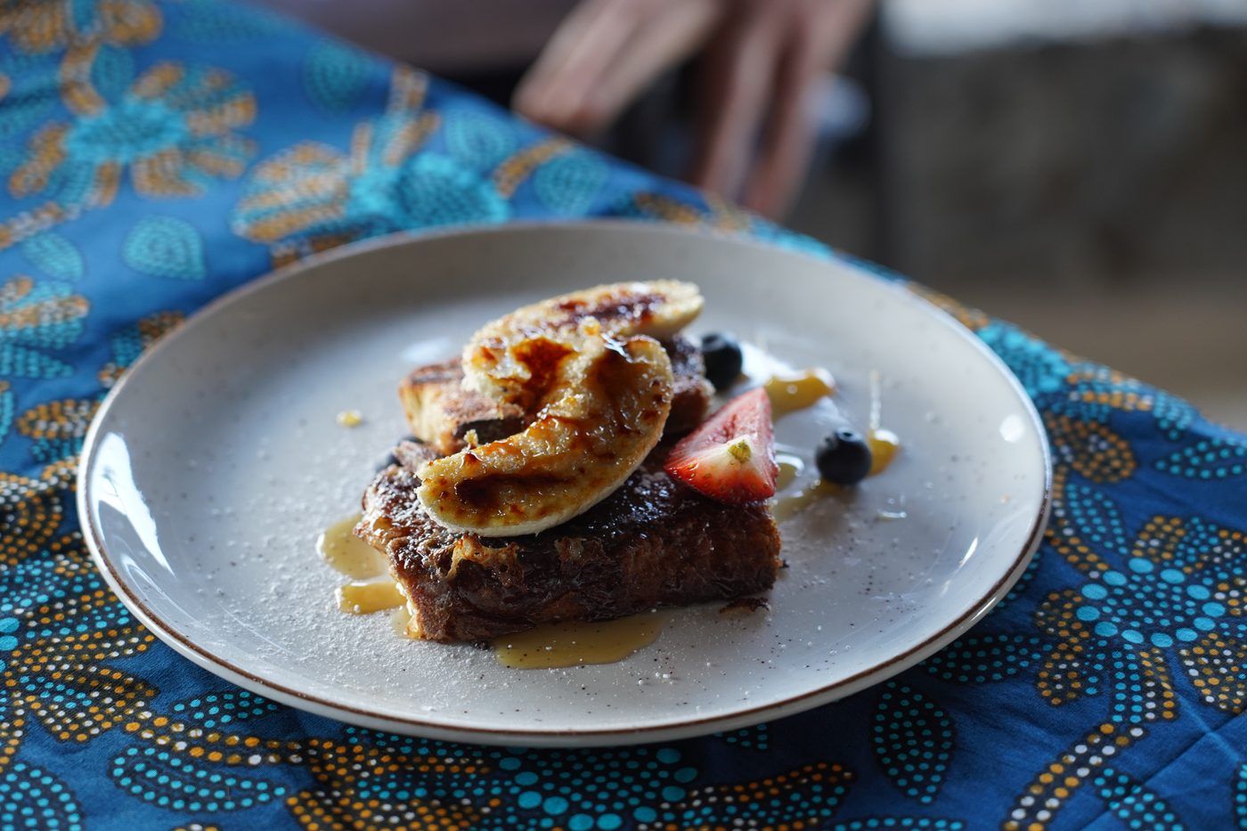 A stack of French toast with bruleed banana, syrup, strawberry and blueberries on a pale plate, set on a blue batik cloth.