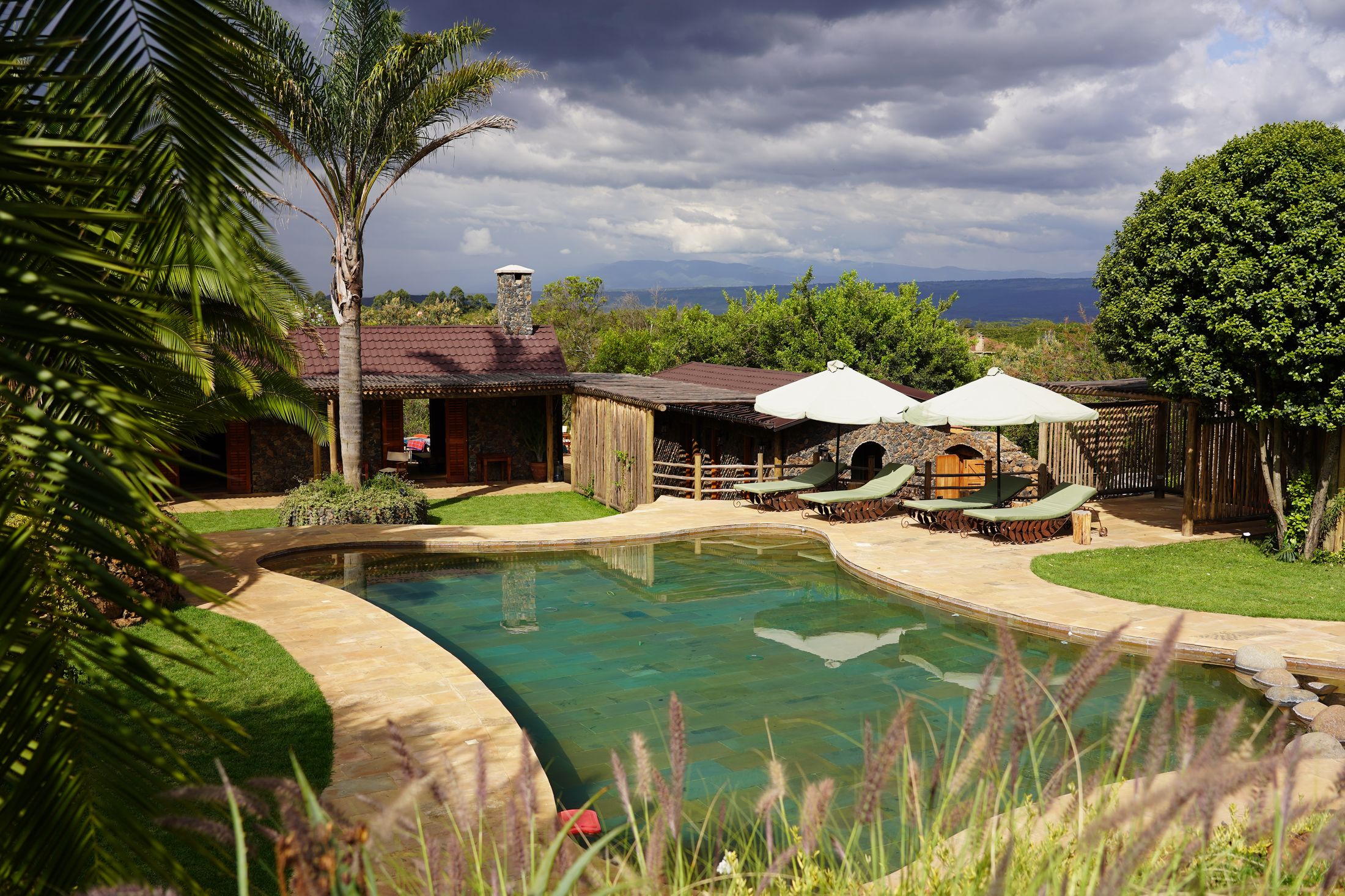 The free-form pool with loungers and parasols under a dramatic storm sky, a palm at the left and hills in the distance.