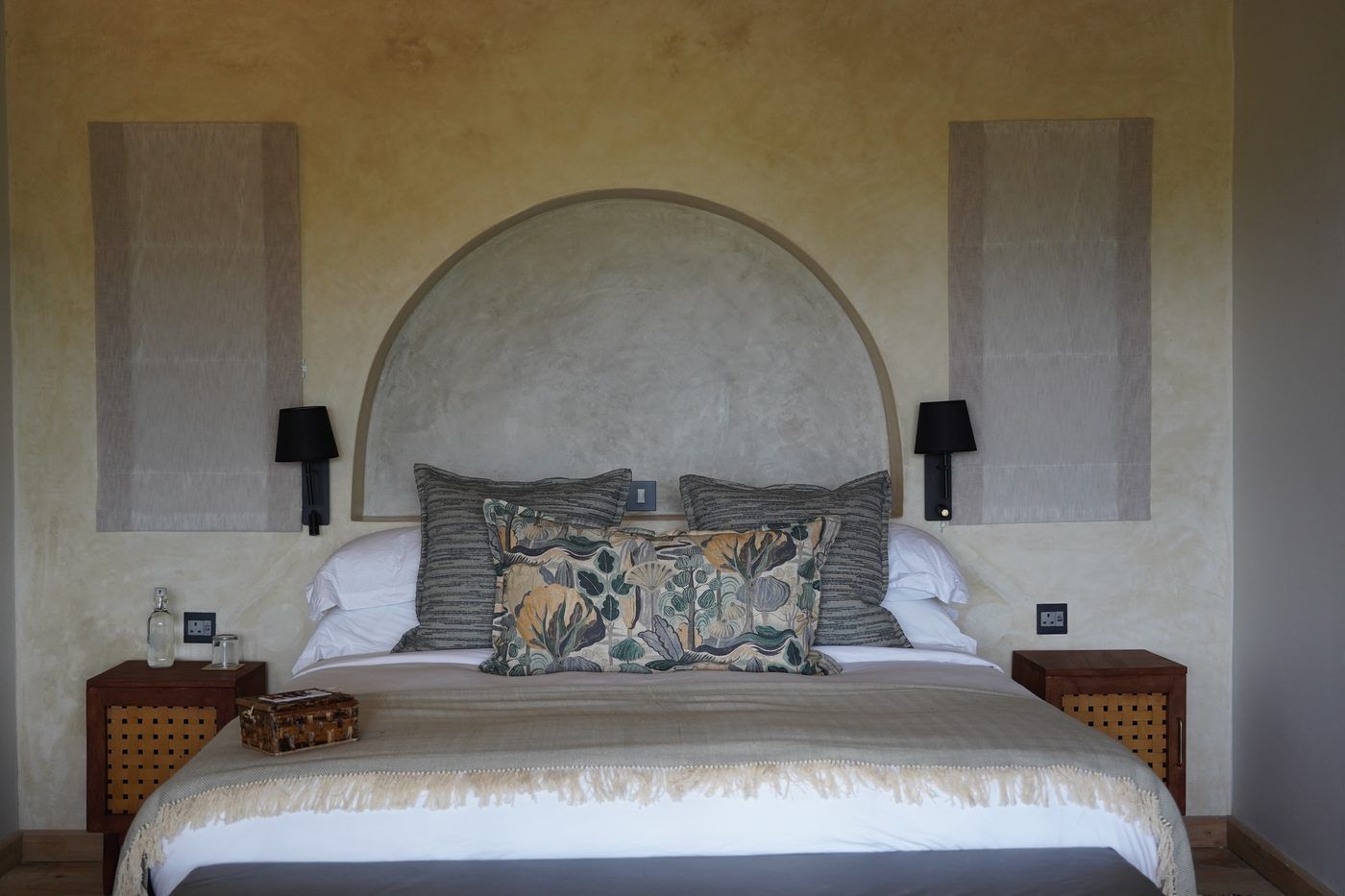 A bed set symmetrically beneath a plaster arch niche on a lime-washed wall at O'Lorien, botanical cushions, black sconces and caned nightstands.