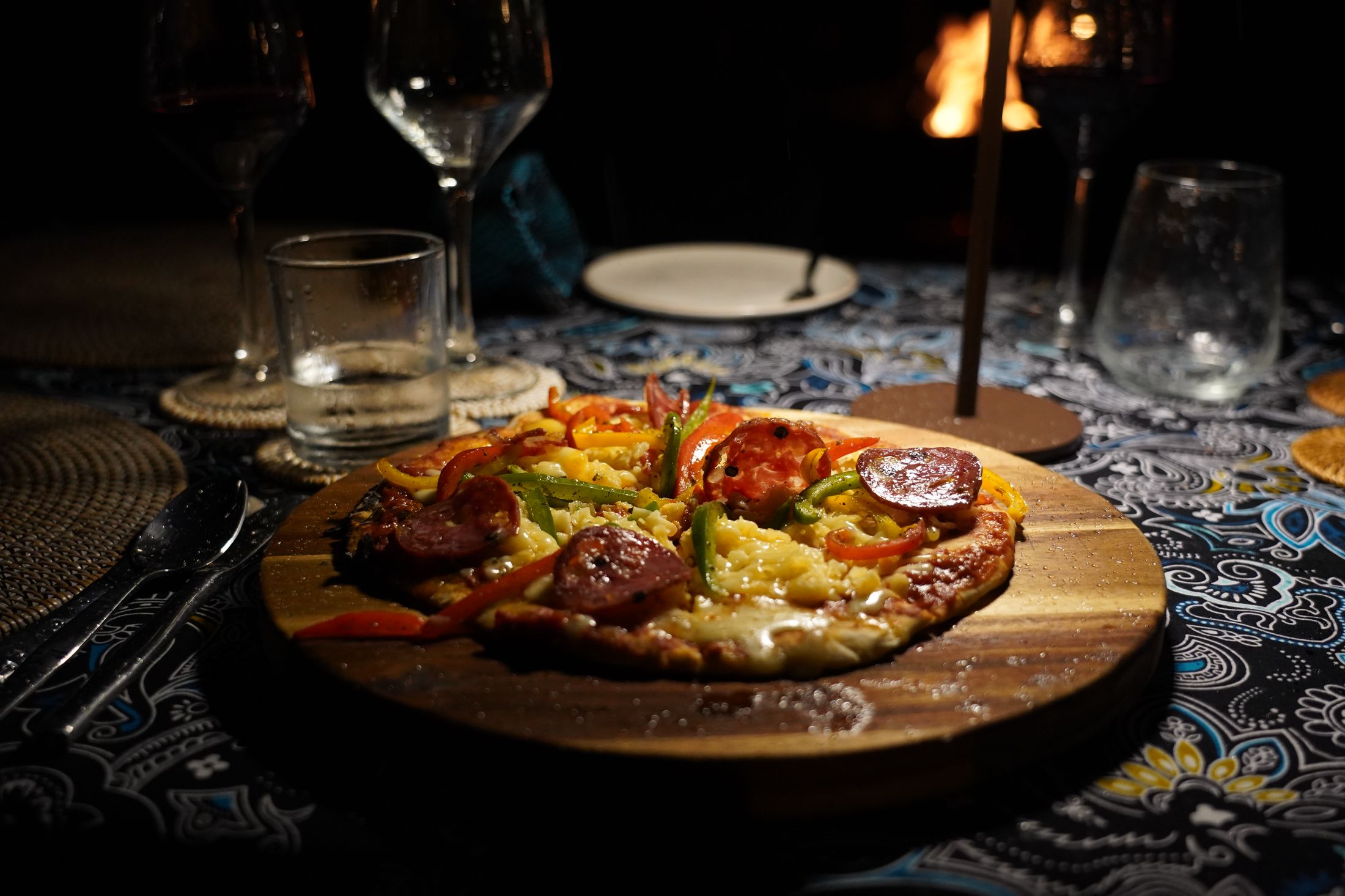 A chorizo and pepper pizza on a wooden board at a dark table, wine glasses around it and the oven's fire glowing behind.