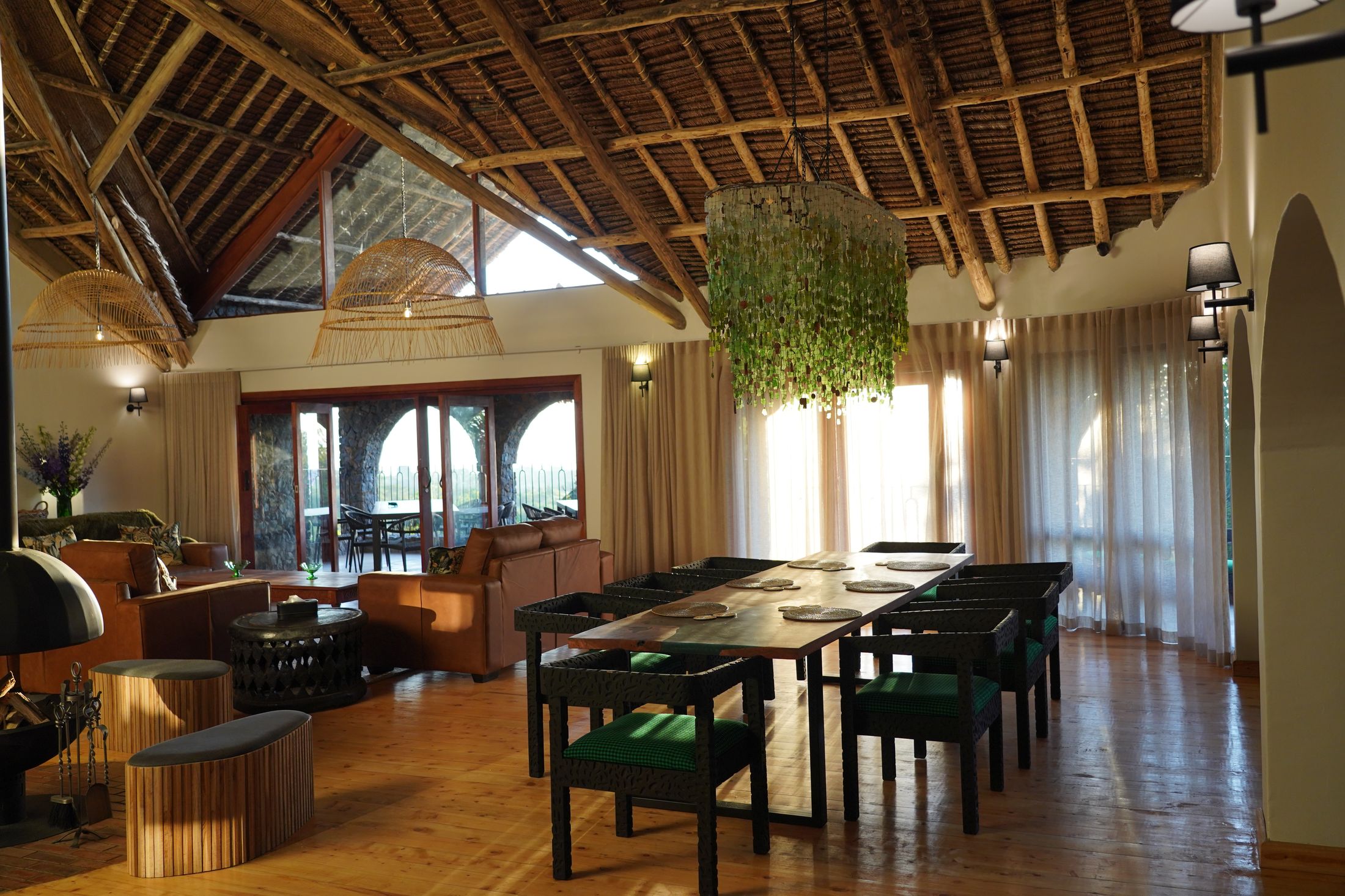 The O'Leleshwa great room — thatch vault, the hood of the suspended cone fireplace at the left edge, the green Malawian-glass chandelier over the long table, low sun through sheer curtains.