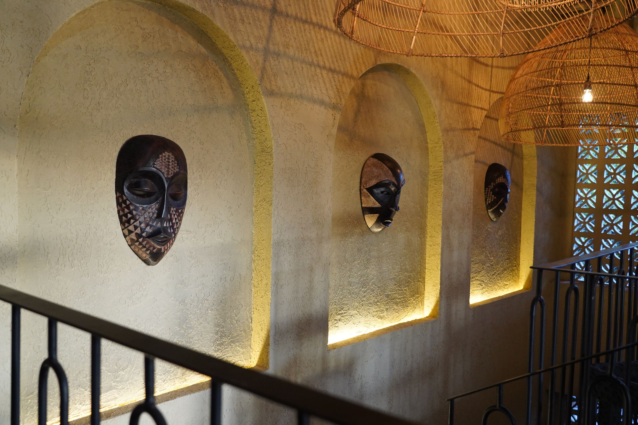 The stair landing at O'Leleshwa — carved masks in underlit arched alcoves, rattan pendants glowing above the iron balustrade.