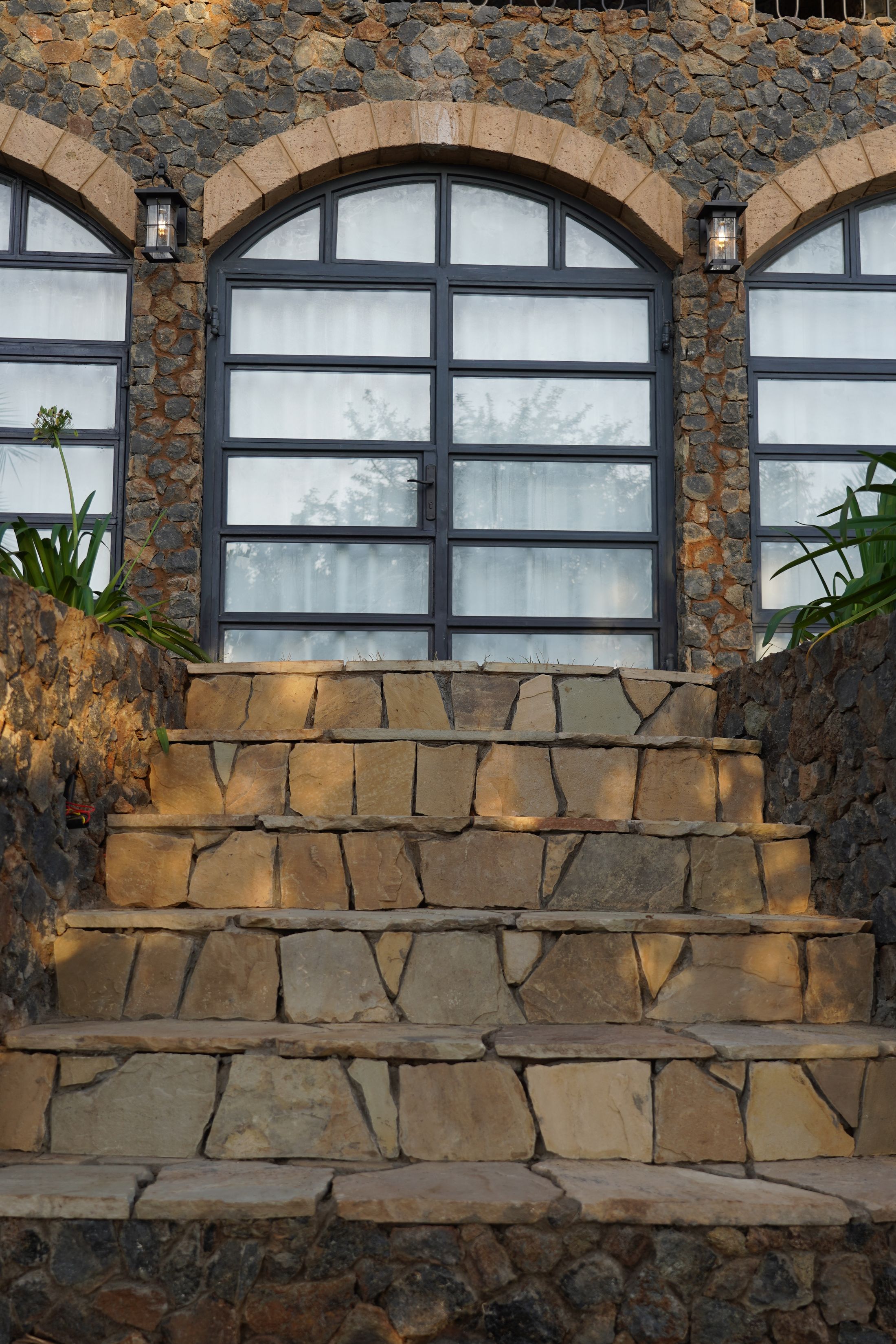 Flagstone steps rising between dry-stone walls to arched steel-framed doors, wall lanterns on either side.