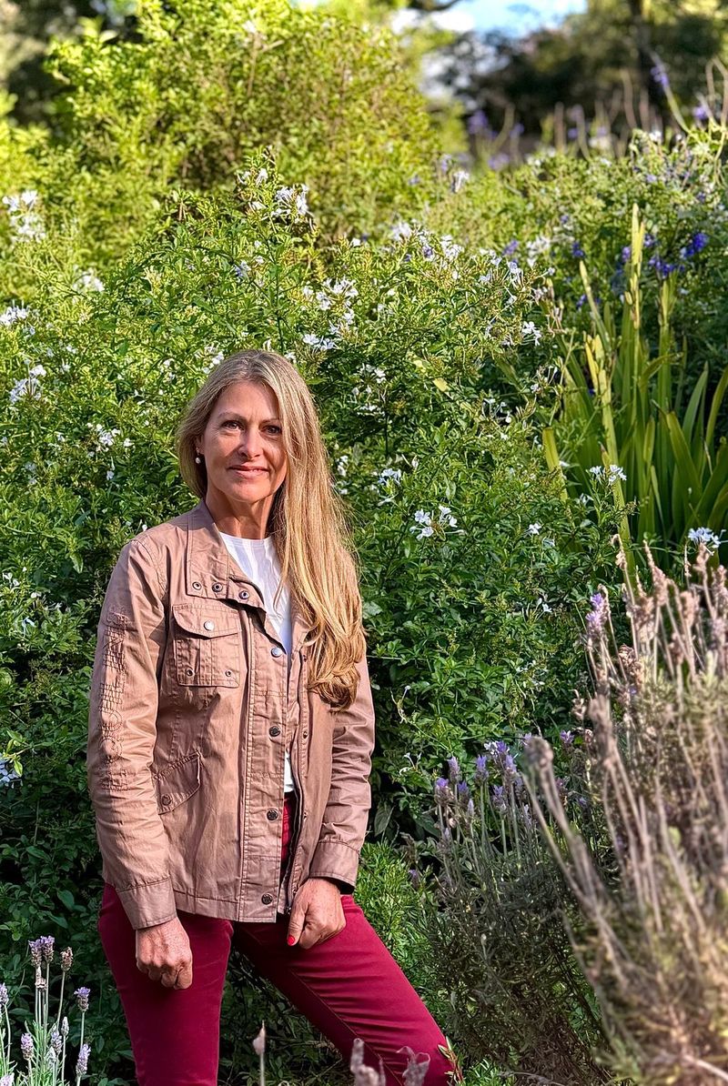 Eileen Neylan in a tan field jacket standing in front of flowering plumbago.