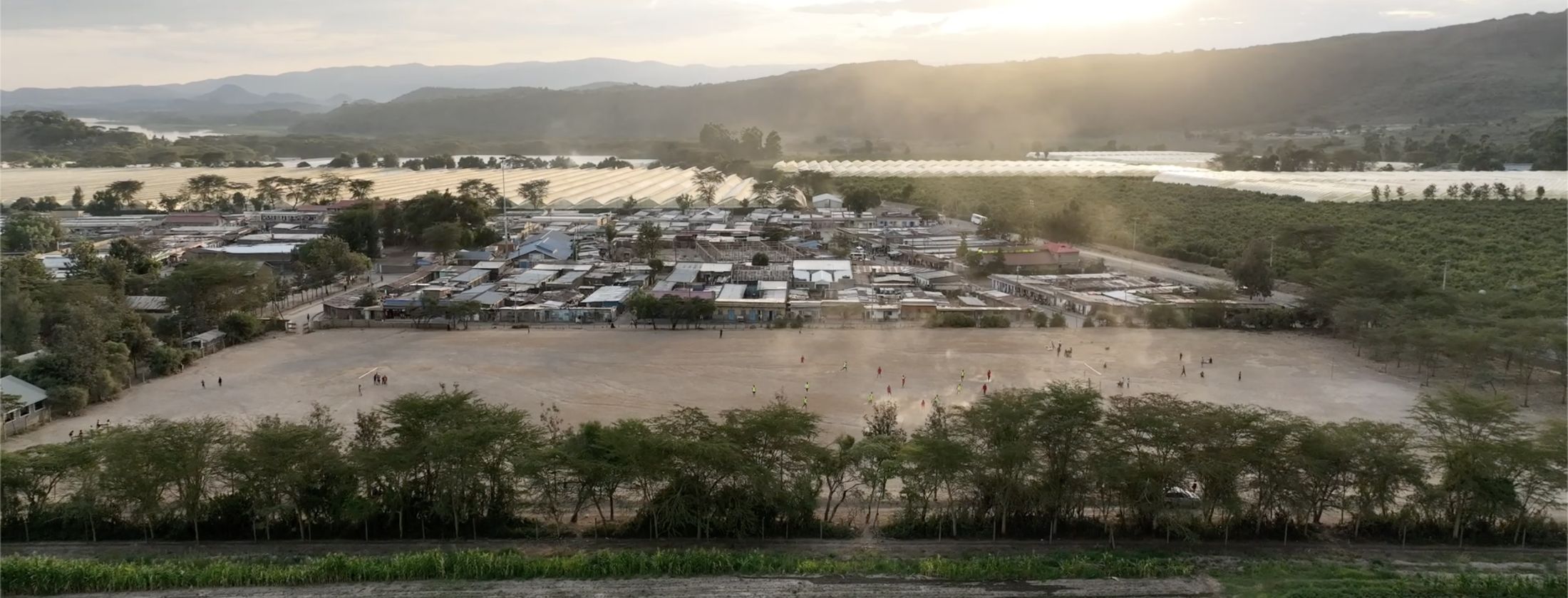 A dusk aerial of Kasarani: a dusty football pitch mid-game, village rooftops, greenhouses and golden haze.