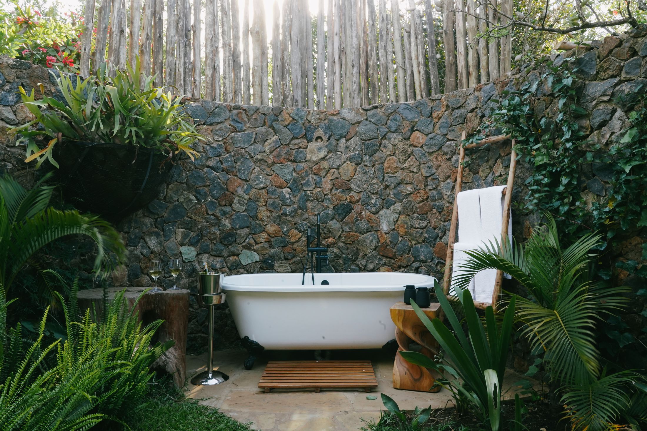 A freestanding bath against the stone wall of O'Lorien's walled garden, ferns around it, the pole fence backlit above.