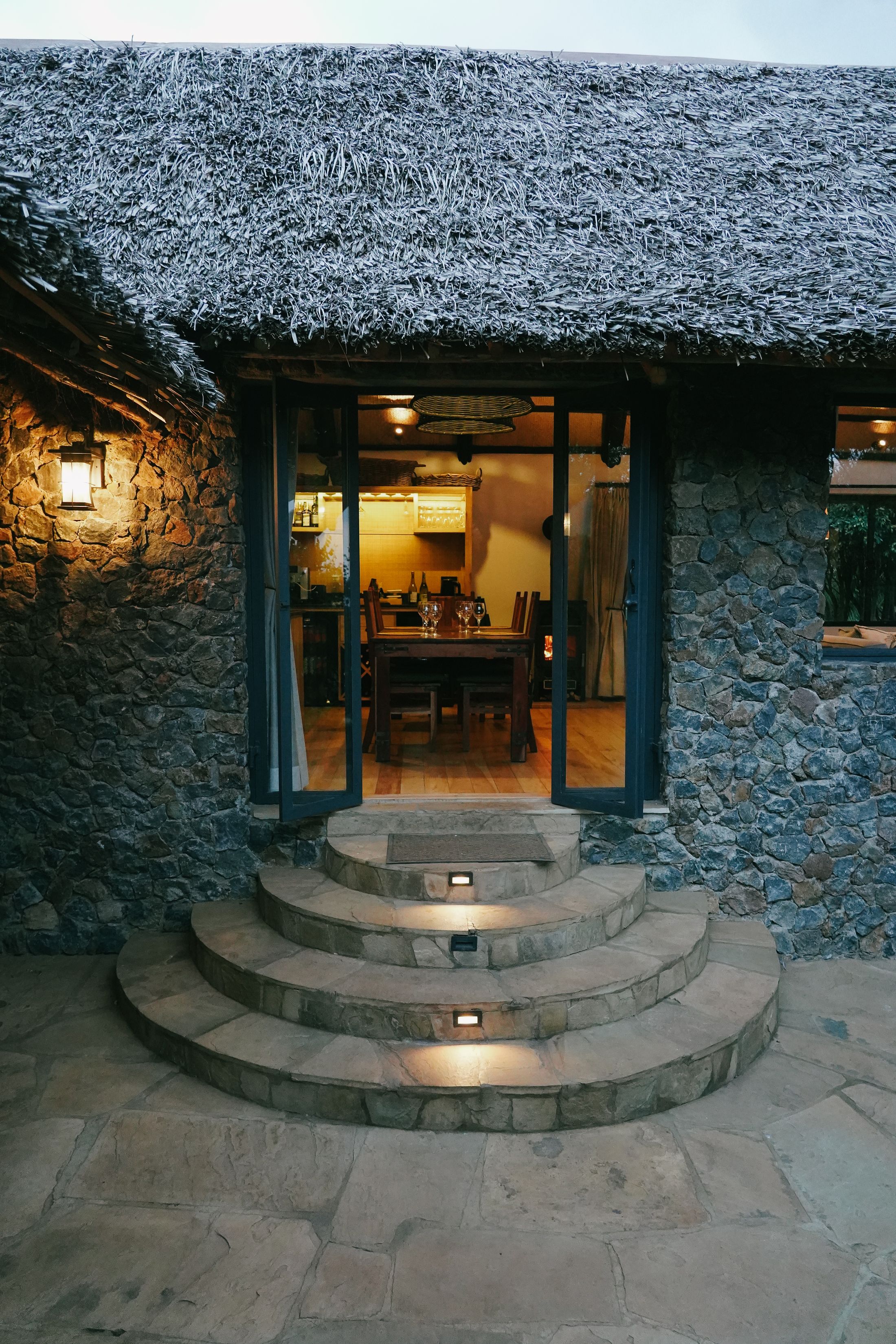 Curved stone steps rising at blue hour to the lamplit kitchen cottage, a table laid inside under thatch.