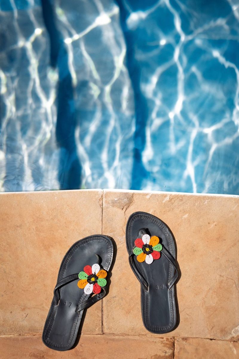 A pair of black leather sandals with beaded flowers on the straps, left on the sandstone coping at the edge of the pool, sunlight breaking on the water above them.