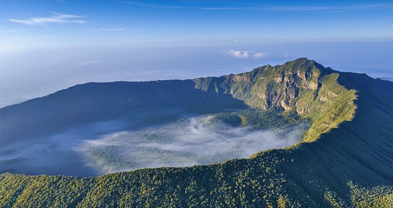 Aerial view of Mount Longonot's crater rim, morning cloud pooled inside the forested caldera under a blue sky.