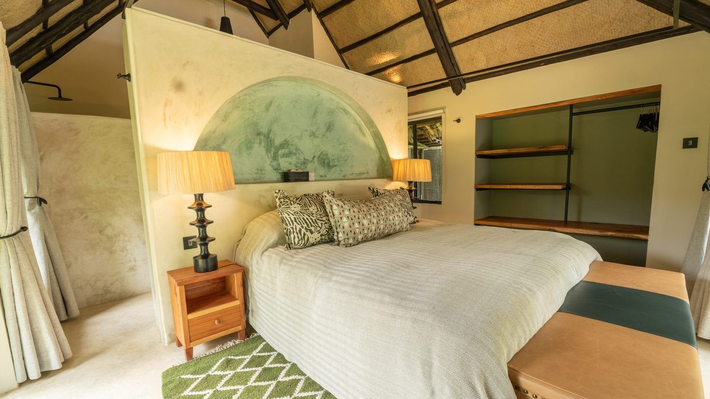 An O'Lorien bedroom beneath thatch on dark poles — a freestanding plaster wall carrying a sage-green fresco arch behind the bed, fringed lamps on carved black stands either side, an open wardrobe niche of live-edge timber shelves to the right, and the shower rose visible on the wall behind the headboard.