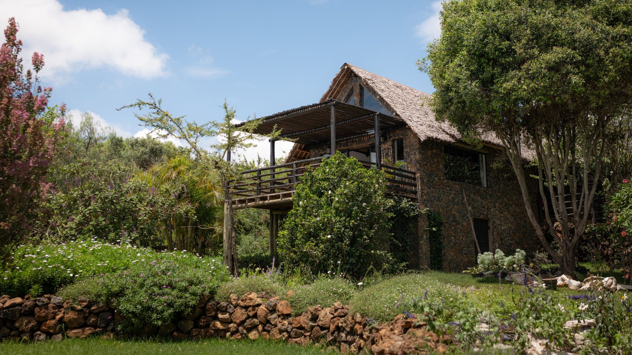 O'Lorien seen across a flowering terraced garden — a two-storey stone house under a thatched A-frame, its pergola deck raised on timber posts among the trees.