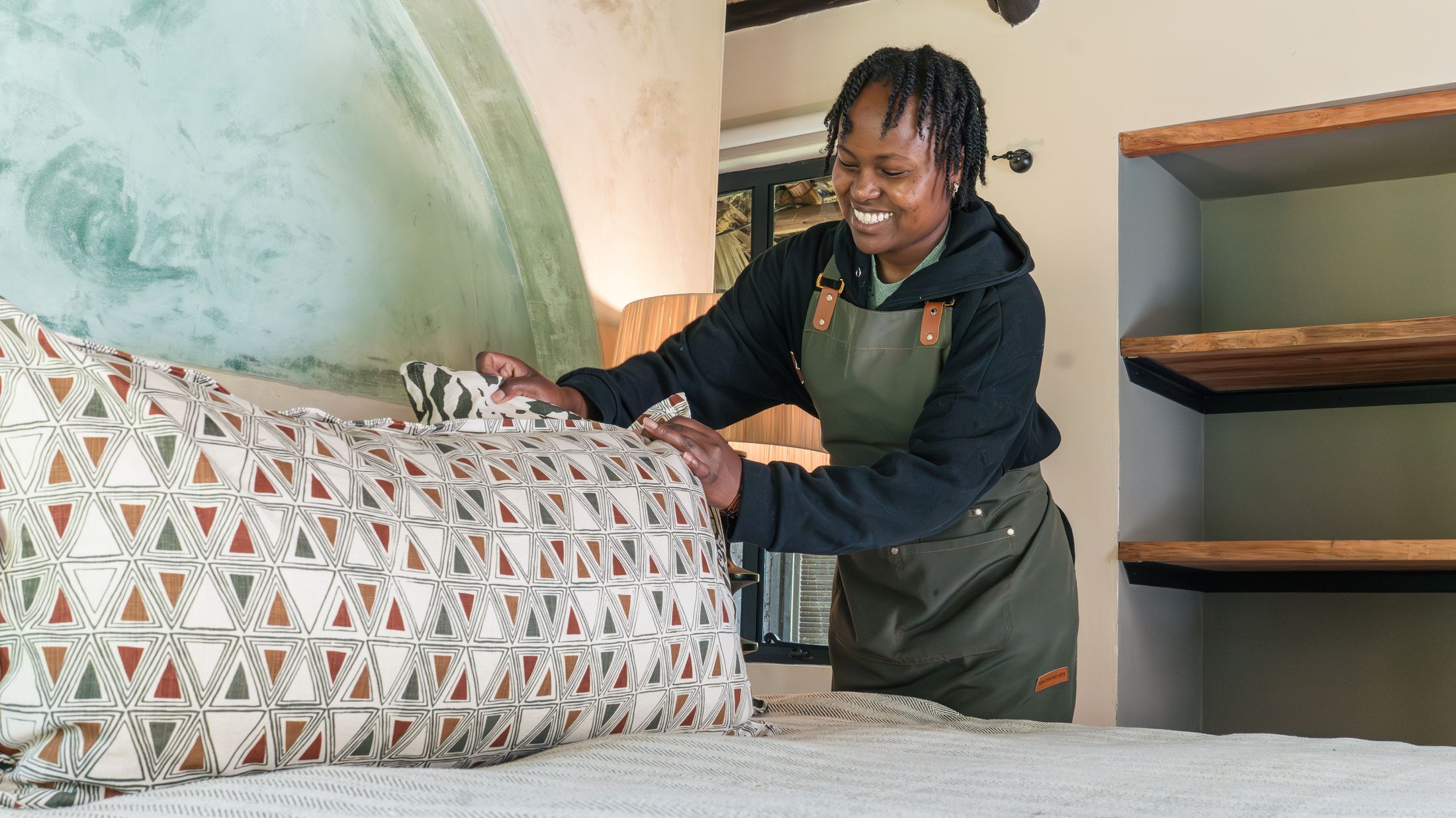 A smiling housekeeper in a green apron dressing a bed with a triangle-print bolster, the green fresco arch behind her.