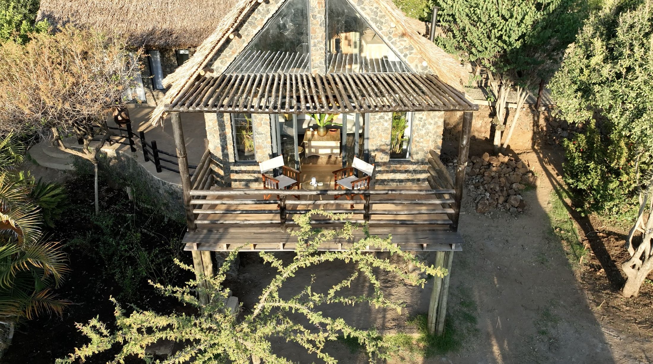 O'Lerai from the air — the pergola deck on gum-pole stilts, two safari chairs at the rail, the glazed triangular gable behind, an acacia spreading over the watering ground below.