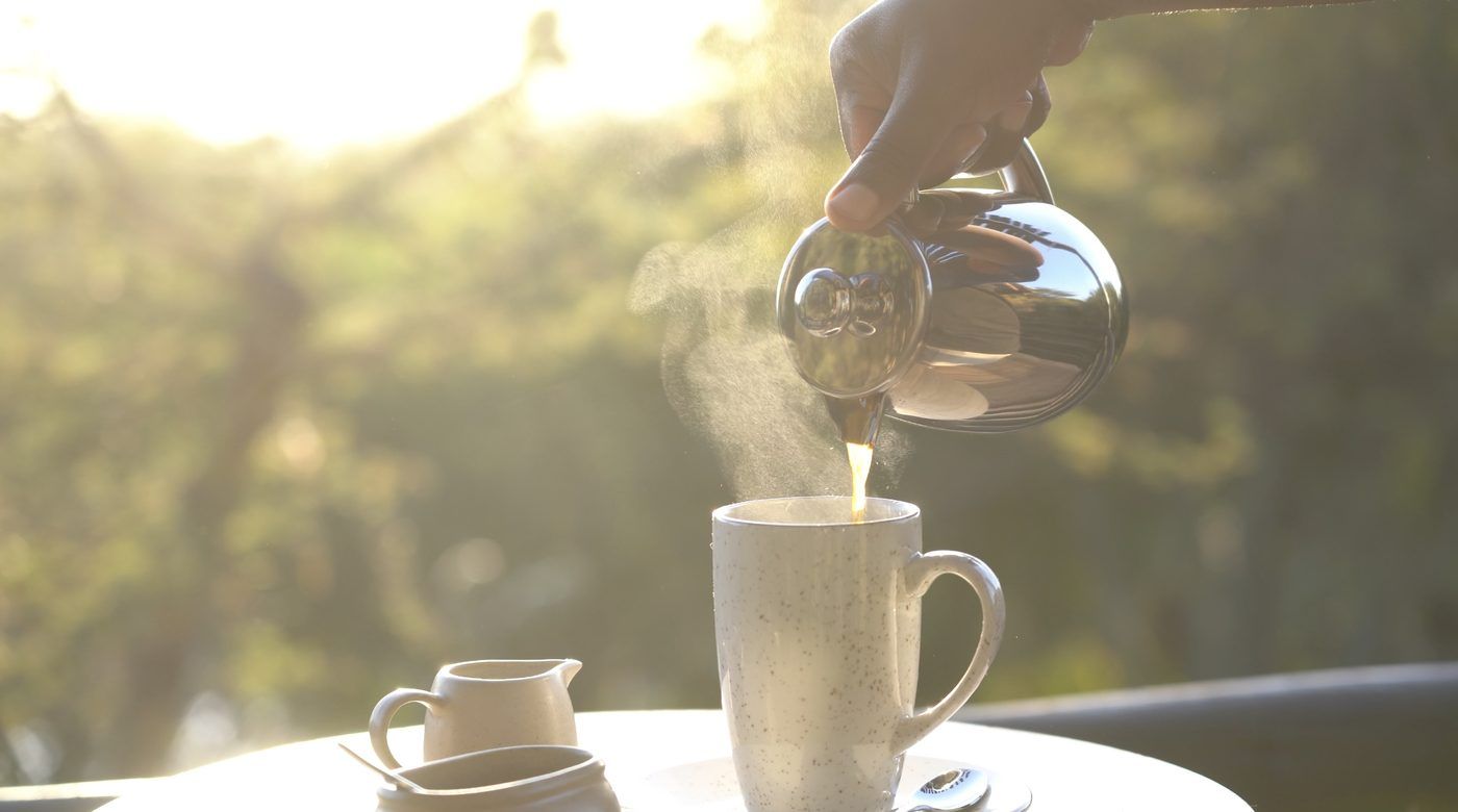 Coffee poured from a steel pot into a speckled mug at sunrise, steam rising, soft garden light behind.